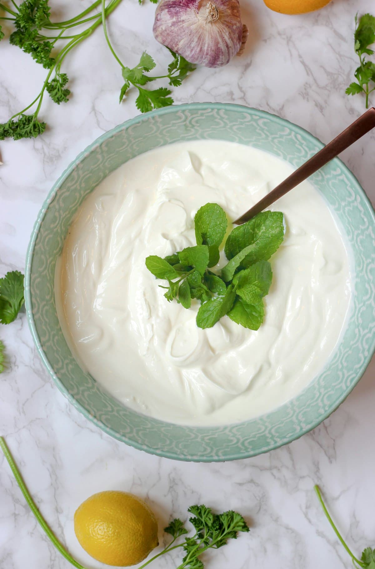 A bowl of thick white yogurt with a spoon, herbs and lemons on a marble counter — taking probiotics with food buffers stomach acid and may improve survival